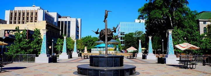 Urban plaza with central fountain statue, blue glass pillars, trees, and city buildings.