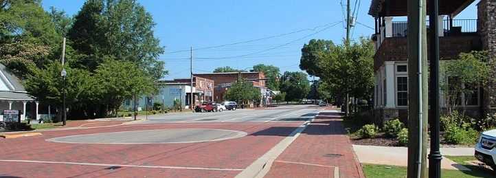 Brick-paved downtown intersection with tree-lined sidewalks, historic storefronts, and a quiet small-town main street.