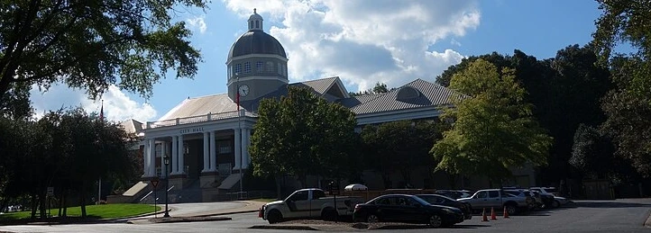 Historic City Hall building with a domed clock tower, surrounded by trees and parked cars under a partly cloudy sky