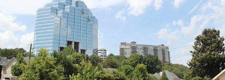Modern glass office tower rising above trees with nearby residential high-rise buildings under a blue sky with scattered clouds.