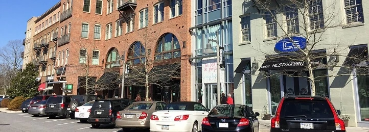 Brick and mixed-use storefront buildings with parked cars lining a downtown street on a clear sunny day.