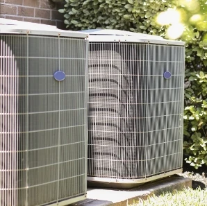 Outdoor HVAC condenser units beside a brick wall and greenery.